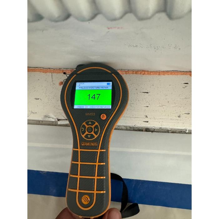 A handheld inspection meter held against an interior wall or trim area, displaying a measurement on its screen.