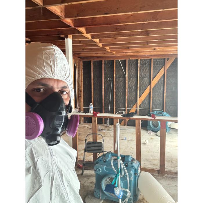 A person in protective gear standing in an unfinished basement or crawlspace with exposed wooden beams and pipes.