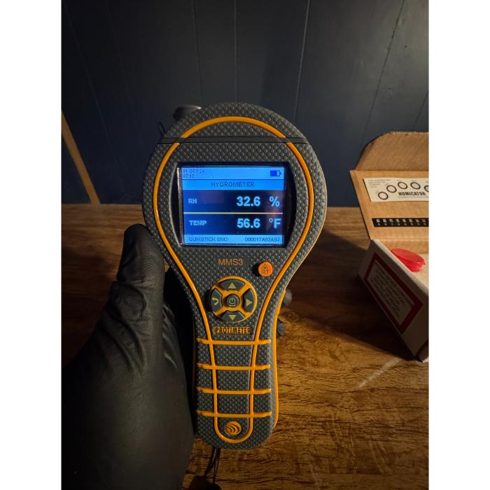 A close-up of a handheld digital inspection meter displaying a reading while held over a wood or laminate floor surface.