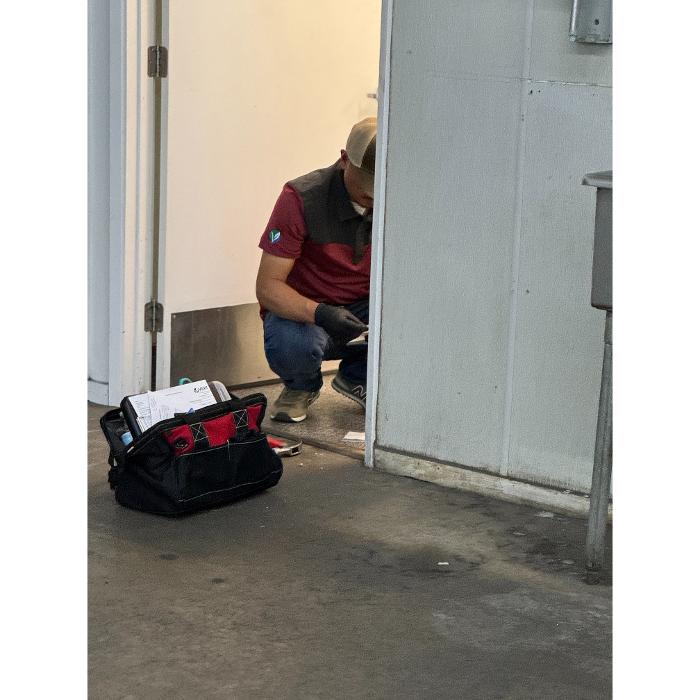A worker kneeling near a doorway with a tool bag on the floor, appearing to inspect or work along the base of the wall.