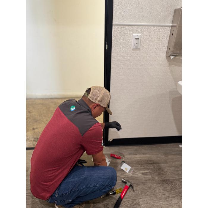 A worker kneeling on the floor inside a room, appearing to inspect or work along the base of a wall.