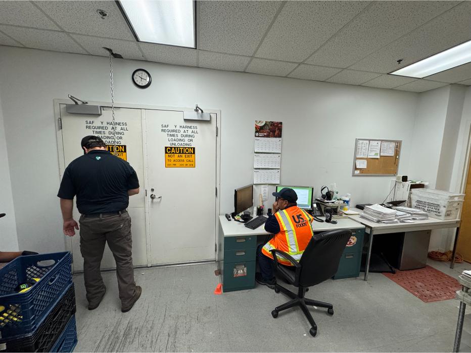 A worker standing in a small office or utility room with desks, chairs, and equipment visible around the space.