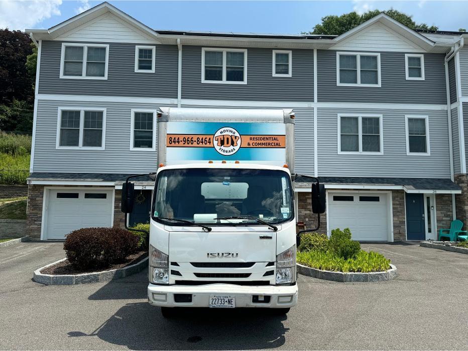 A branded TDY Moving & Storage box truck parked in the driveway of a modern multi-unit townhouse and condominium complex.