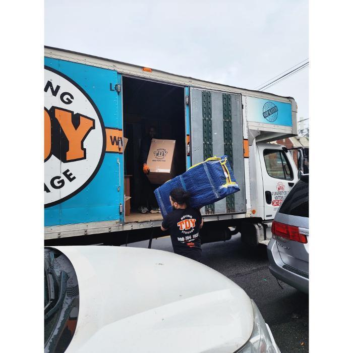 A TDY Moving and Storage professional carefully loading a heavily padded and wrapped blue piece of furniture into the back of a moving truck filled with moving boxes.