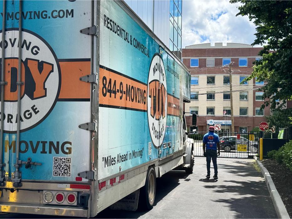 A professional mover walking toward a parked TDY Moving and Storage box truck outside a large commercial building and apartment complex in New York.