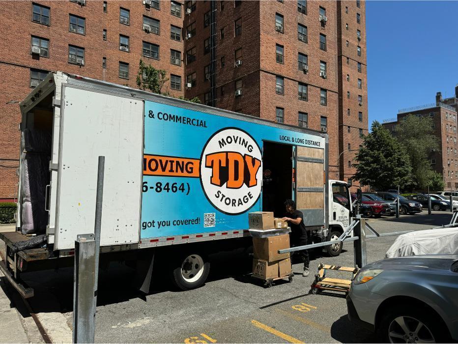 A blue and orange TDY Moving & Storage truck with its ramp deployed, parked on a busy Brooklyn street outside a tall brick residential apartment building.