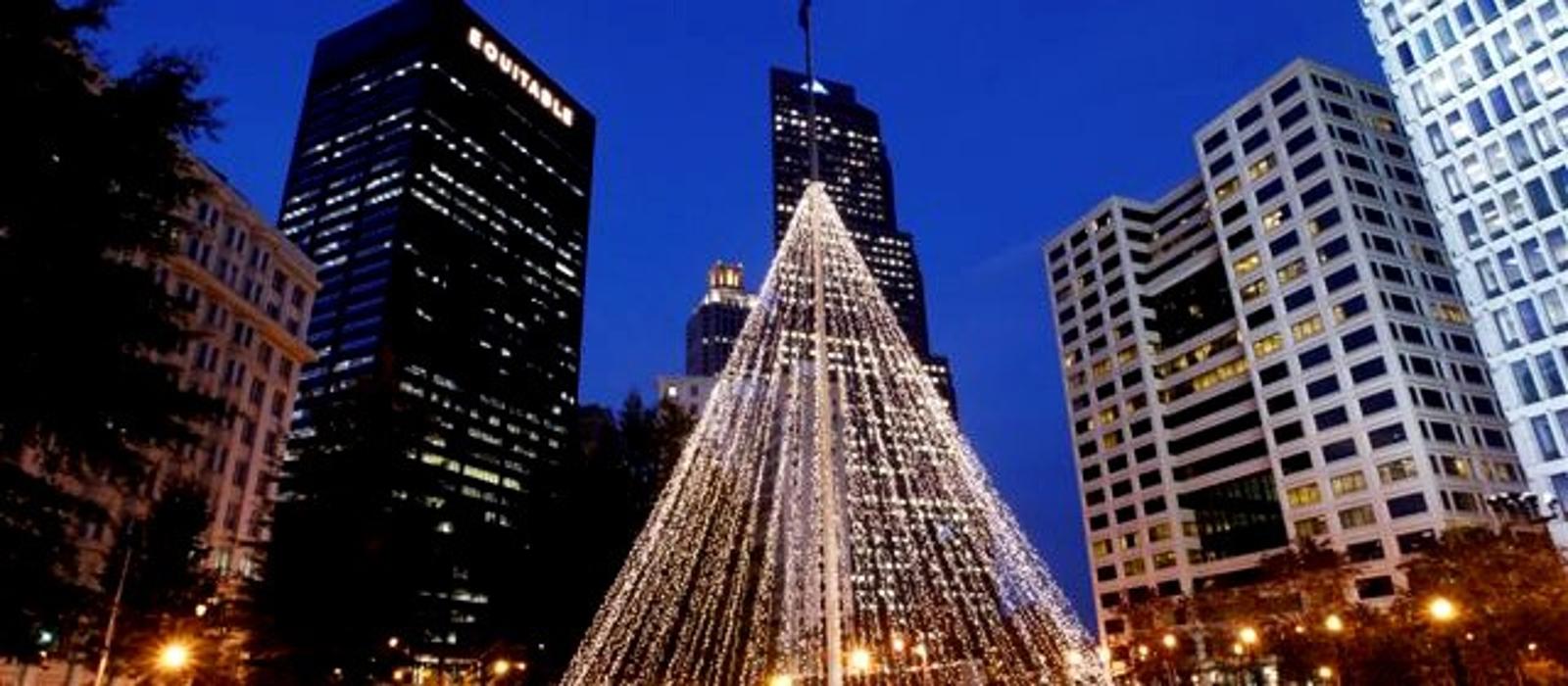 A large illuminated Christmas tree display surrounded by tall buildings, creating a striking holiday scene in an urban plaza at dusk.