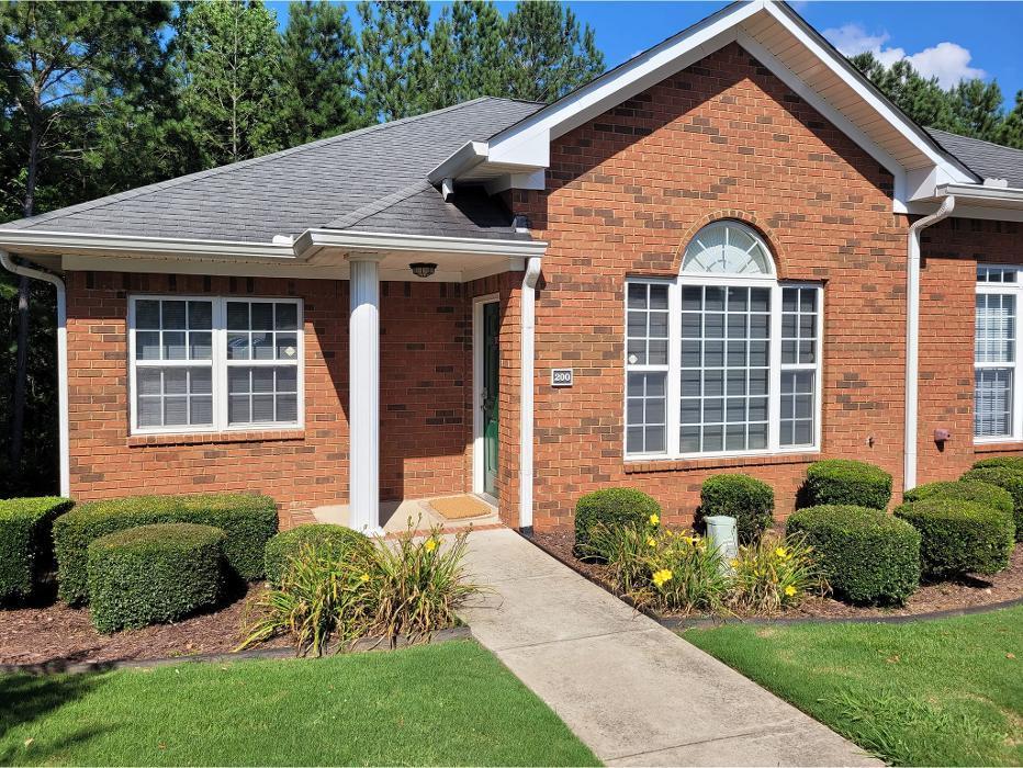 Red brick single-story residential building with gray sloped roof, white trim, large front windows, arched window detail, concrete walkway, landscaped bushes and plants in front yard, tall trees in background.