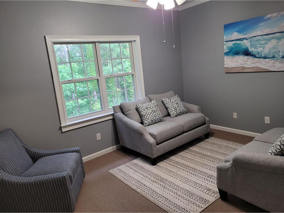 Interior room with light gray walls, large multi-pane window overlooking green foliage, gray upholstered sofa with patterned pillows, striped accent chair, area rug, framed artwork on wall showing ocean wave, minimal furnishings, natural light.