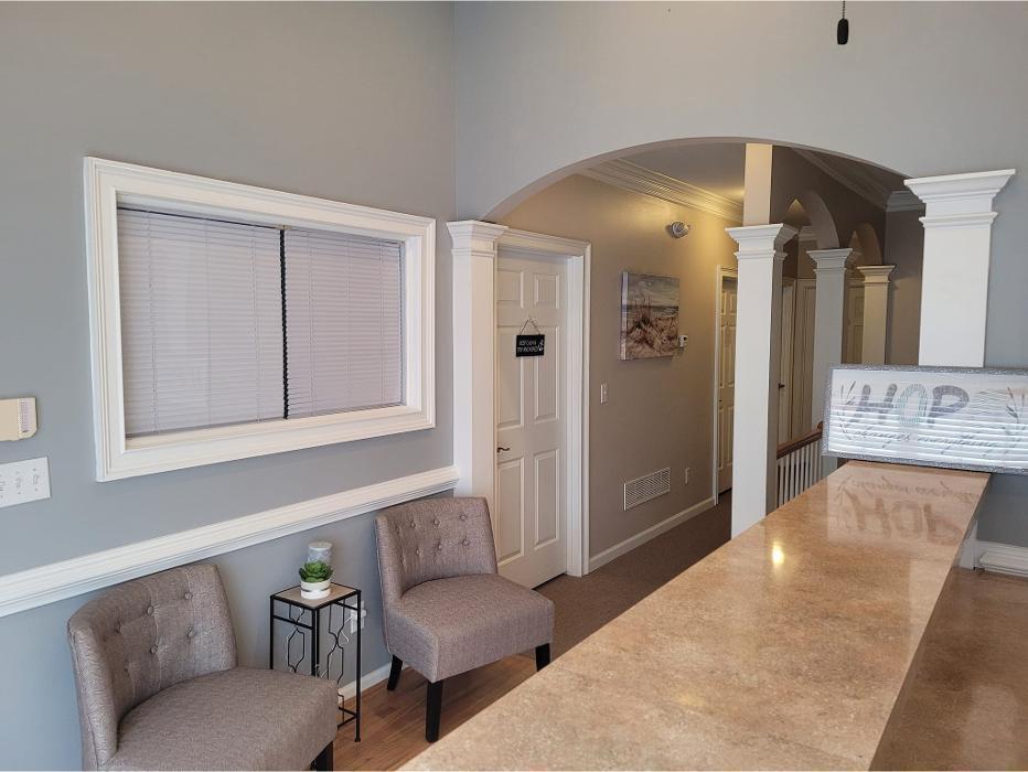 Interior reception or waiting area with light gray walls, tan/beige colored chairs, wooden reception counter, window with blinds, cream-colored doorways and archways with white trim, hardwood flooring, small side table with plant.