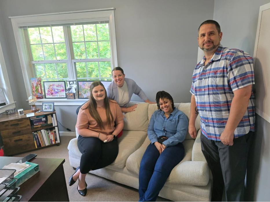 Five people posing together indoors in front of large multi-pane window with natural light, casual clothing, standing and sitting on cream-colored sofa, bookshelves visible in background, professional yet friendly group setting.