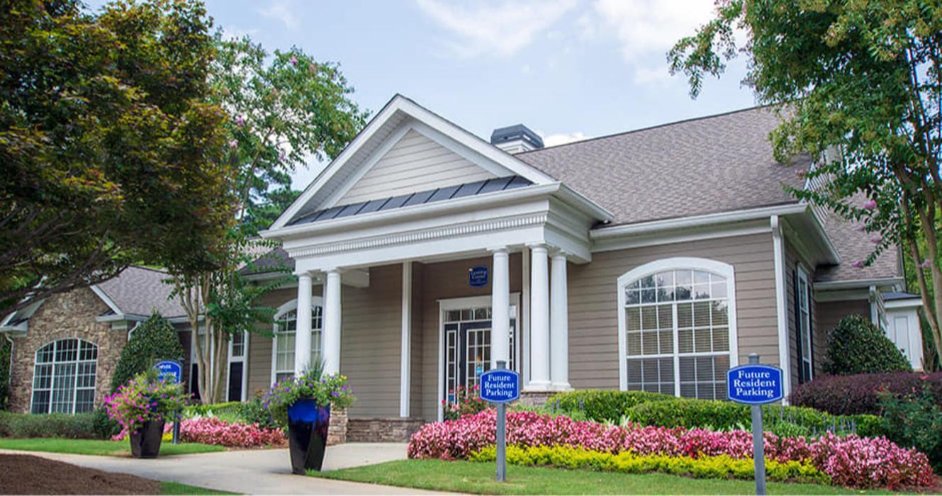 Tan/beige single-story building with white columns, pitched roof, brick accent wall visible on left side, colorful flowering plants (pink, yellow blooms) in landscaping, "Future Resident Parking" signs visible, mature trees framing sides.