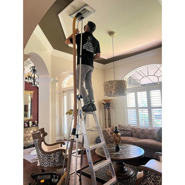HVAC professional on a ladder cleaning an overhead air vent in a residential living space as part of an air duct cleaning service.