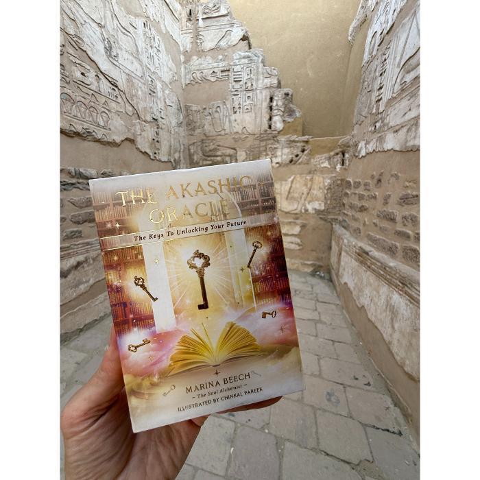 The Akashic Oracle card deck box by Marina Beech, The Soul Alchemist, held up against ancient stone walls with hieroglyphics