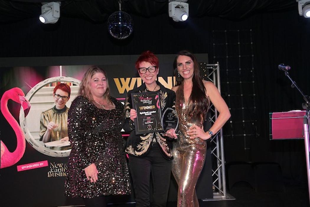 Marina Beech on stage at the Flamingo Business Awards receiving her Spiritual Leader Award, flanked by two women, with the winner screen visible behind them