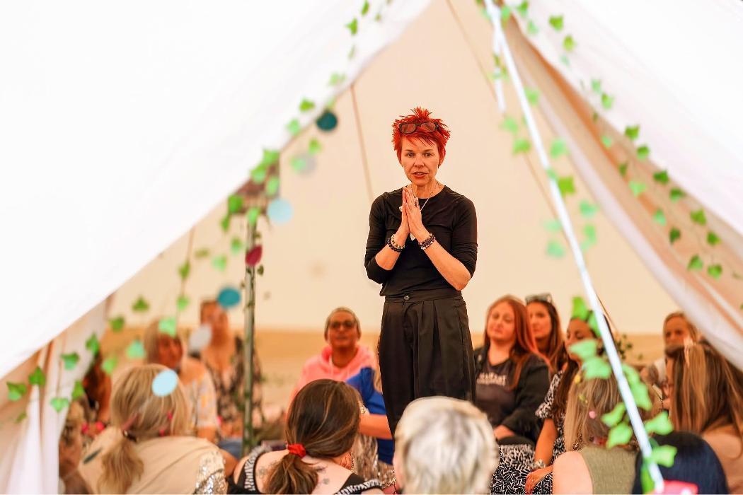 Marina Beech speaking to a seated audience inside a large bell tent at an outdoor event, hands in prayer position