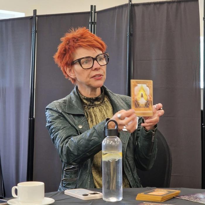 Marina Beech holding up an Akashic Oracle Card at an exhibition or event, seated at a table with oracle cards and her water bottle