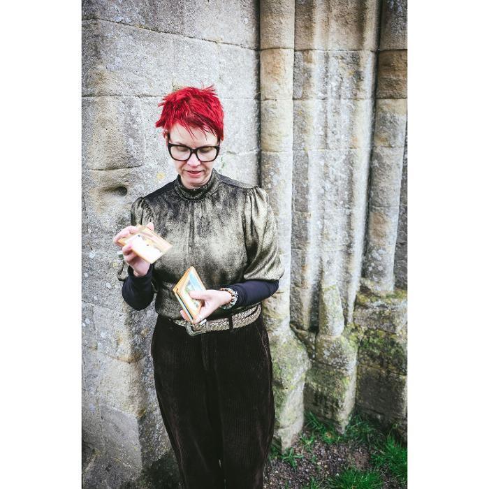 Marina Beech reading Akashic Oracle Cards against an ancient stone wall, wearing a gold metallic top