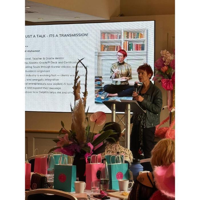 Marina Beech speaking on stage at a live event, holding a microphone, with a large screen behind her showing her presentation slide titled "Not Just a Talk, It's a Transmission"