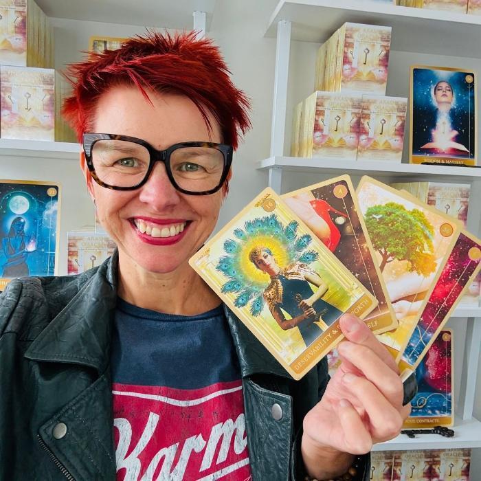Marina Beech smiling and holding a fan of Akashic Oracle Cards, with shelves of Akashic Oracle Card decks displayed behind her