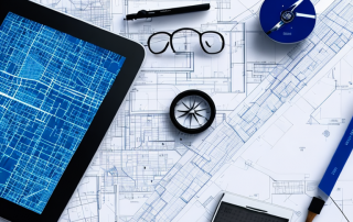 Architectural blueprints on table with ruler, compass, and tablet displaying zoning maps.