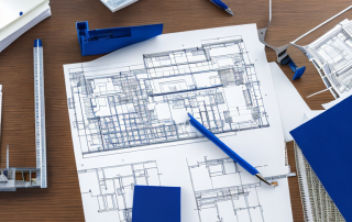 gut organisierter Architekturschreibtisch mit Bauplaenen, Modellbau und Zeichengeraeten, Haende zeigen auf Bauplaene, Zusammenarbeit in der Stadtplanung.