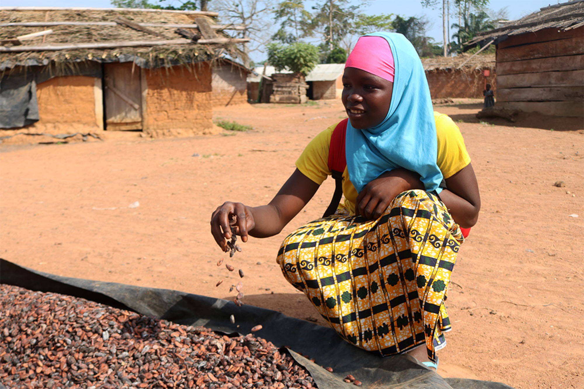 Young woman in colorful clothing sorting dried cocoa beans in a rural village, with thatched-roof huts in the background.